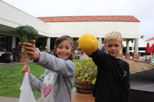Children holding up fresh grapefruit and broccoli at a farmers market for WaveCrest Cafe
